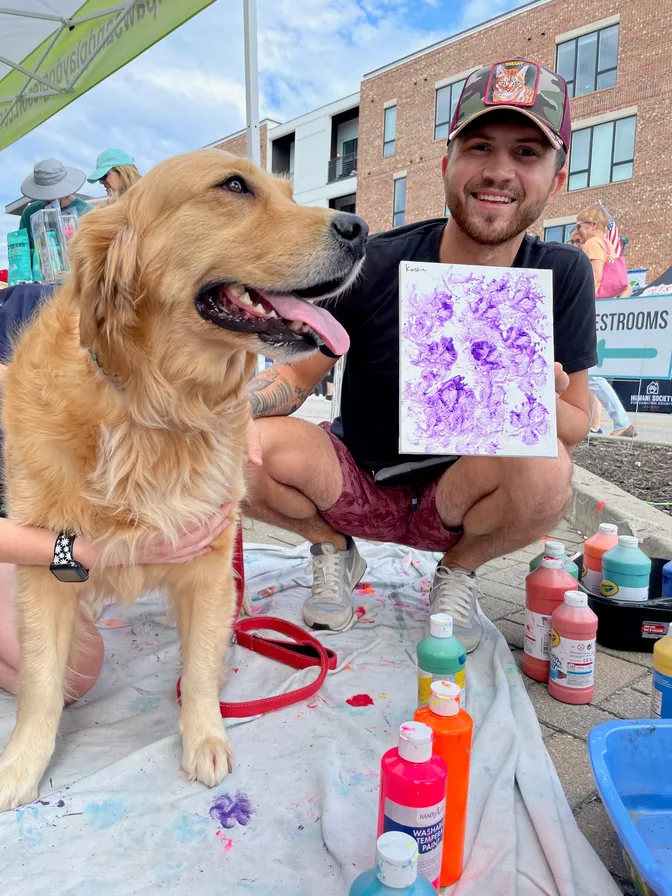 Skyler Braun crouching beside a golden retriever, holding up a paw-print painting at a Humane Society for Hamilton County event